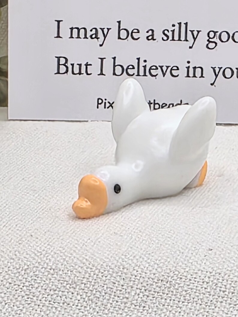 May include: A white ceramic goose figurine with an orange beak and feet. The goose is lying down with its head tilted. Text on a card behind the goose reads, "I may be a silly goose, but I believe in you."