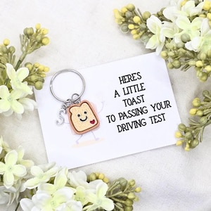 Peut inclure: Un porte-clés fantaisie avec un charm en forme de toast souriant et un cœur, attaché à un porte-clés argenté avec un charm initial. La carte indique : "Voici un petit toast pour avoir réussi votre permis de conduire".