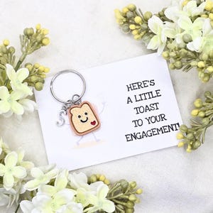 May include: A novelty keychain featuring a cartoon toast charm with a smiling face and heart. The keychain includes a silver initial charm and is presented with a card that reads, "Here's a little toast to your engagement!"