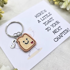 Könnte beinhalten: Ein Schlüsselanhänger mit einem Toast-Charm mit lächelndem Gesicht und Herz, plus einem silbernen Buchstaben "S". Die Karte lautet: "Here's a little toast to your next chapter!"