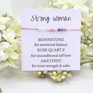 May include: A white card with the text "Strong woman" and a description of the stones in a bracelet: Moonstone for emotional balance, Rose Quartz for unconditional self-love, Amethyst for inner strength & calm.