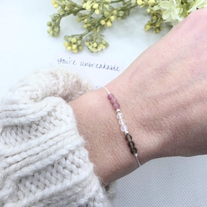 May include: A delicate bracelet with a white string and small pink, clear, and brown beads. The bracelet is on a person's wrist and the text "you're unbreakable" is visible in the background.