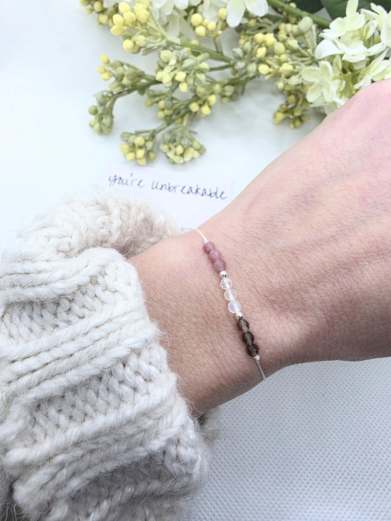 May include: A delicate bracelet with a white string and small pink, clear, and brown beads. The bracelet is on a person's wrist and the text "you're unbreakable" is visible in the background.
