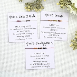 May include: Three cards with the words "you're unbreakable", "you're enough", and "you're unstoppable" printed on them. Each card features a string bracelet with different colored gemstones and a description of the stones' meanings.