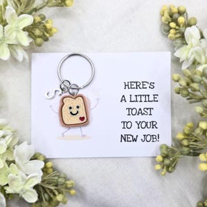May include: A novelty keychain featuring a smiling toast slice with a heart, attached to a silver keyring. The card reads "Here's a little toast to your new job!" surrounded by white and green floral accents.