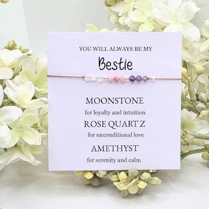 May include: A friendship bracelet with a string of clear, pink, and purple beads. The card reads "YOU WILL ALWAYS BE MY Bestie" and lists the properties of the gemstones: Moonstone, Rose Quartz, and Amethyst. White flowers are in the background.