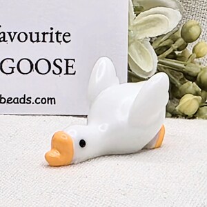 May include: A small, white ceramic goose figurine with an orange beak and feet. The goose is lying down with its wings slightly spread. The text "favourite GOOSE" is visible in the background.