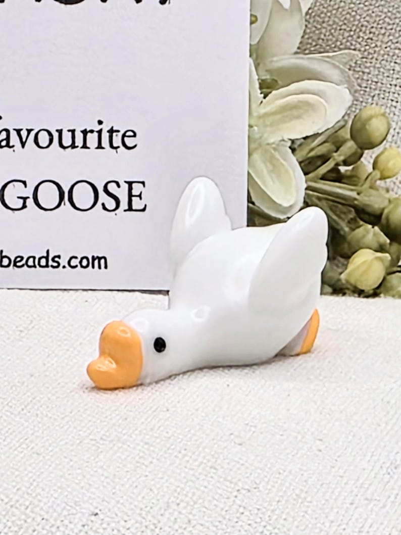 May include: A small, white ceramic goose figurine with an orange beak and feet. The goose is lying down with its wings slightly spread. The text "favourite GOOSE" is visible in the background.