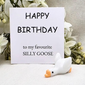 May include: A white card with the words "HAPPY BIRTHDAY" and "to my favourite SILLY GOOSE" in black text. A small white goose figurine with an orange beak and feet is in front of the card. The background has white flowers.