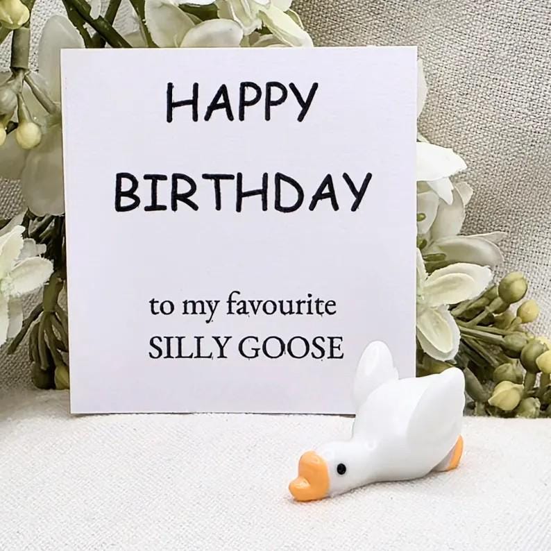 May include: A white card with the words "HAPPY BIRTHDAY" and "to my favourite SILLY GOOSE" in black text. A small white goose figurine with an orange beak and feet is in front of the card. The background has white flowers.