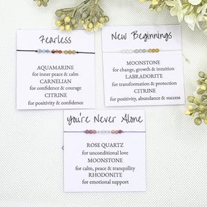 May include: Three cards with the words "Fearless", "New Beginnings", and "You're Never Alone" printed on them. Each card features a bracelet with different colored gemstones and a description of the stones' meanings.