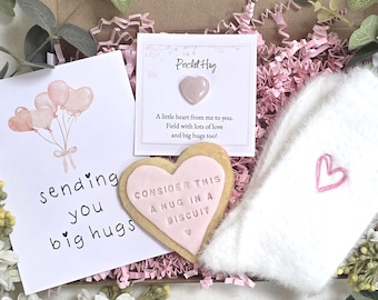 May include: A gift box containing a greeting card with pink heart balloons and the text sending you big hugs, a Pocket Hug card with a small heart, a heart-shaped biscuit with the text CONSIDER THIS A HUG IN A BISCUIT, and white fuzzy socks.