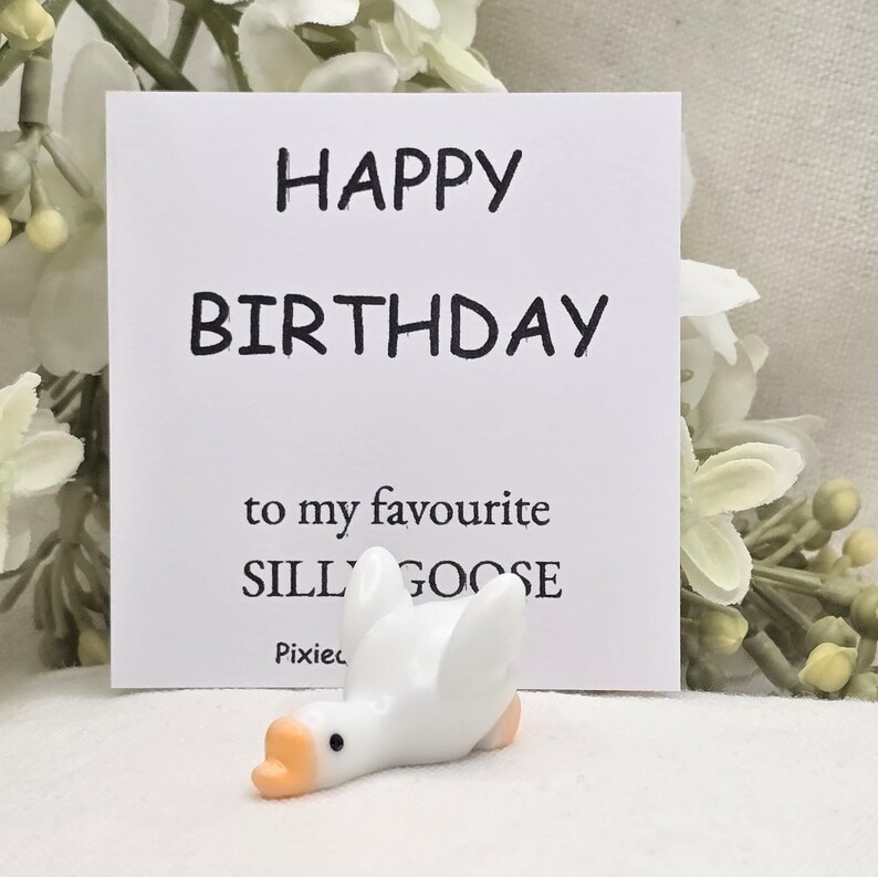May include: A white ceramic goose figurine with an orange beak and feet, lying down in front of a white card. The card reads "HAPPY BIRTHDAY to my favourite SILLY GOOSE".