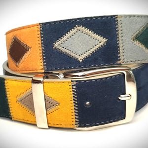 Op de afbeelding: Een kleurrijke riem met een patchwork-ontwerp. De riem heeft delen van oranje, gele, marineblauwe en grijze suède, elk met een gestikt ruitpatroon. Een zilverkleurige gesp zet de riem vast.