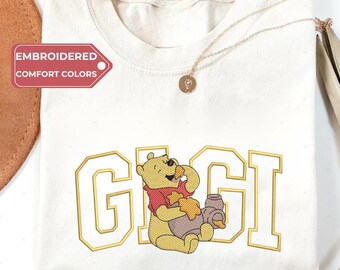 May include: Cream-colored t-shirt with the word "GIGI" in gold lettering and a cartoon bear illustration. The bear is wearing a red scarf and holding a honey pot. The text "EMBROIDERED COMFORT COLORS" is on a pink tag.