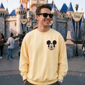 A man wearing a yellow sweatshirt with a Mickey Mouse logo stands in front of a Disney castle.