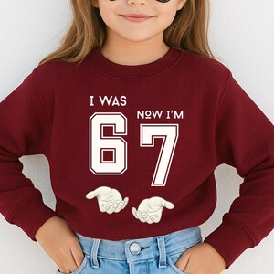 A young girl wearing a red sweater with the text "I WAS 6 NOW I'M 7" printed on it.