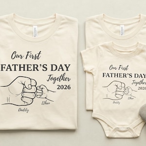 May include: Cream-colored matching t-shirts and a onesie with the text "Our First Father's Day Together 2026" and a graphic of two fists bumping. The onesie has a pair of gray baby shoes next to it. Family apparel.