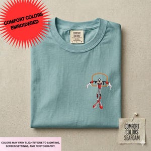 a light blue t-shirt with a graphic of a character wearing a red and white outfit, standing in a basketball hoop. The t-shirt is displayed on a flat surface, and there is a tag attached to it.
