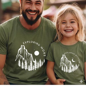 May include: Two olive green t-shirts with matching mountain and forest designs. One shirt says "EXPLORING MORE" and the other says "EXPLORING WITH DAD". The shirts are worn by a smiling adult and a child.