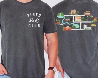 May include: Dark gray t-shirt with the text "TIRED DADS CLUB" in white on the front. The back of the shirt features a colorful illustrated map with buildings, trees, and a river. The shirt is made of a soft, comfortable material.