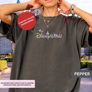 A person wearing a gray t-shirt with the Disney World logo, posing with their hands on their ears.