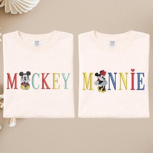 two white t-shirts with the word "MACKKEY" and the name "MANNIE" printed on them, along with illustrations of Mickey Mouse and Minnie Mouse.