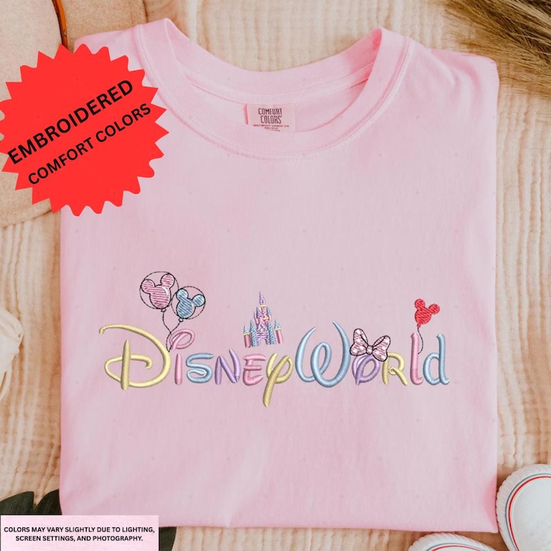 A pink t-shirt with the Disney World logo embroidered on it, along with various Disney-themed characters and landmarks such as Mickey Mouse, Minnie Mouse, and the castle.