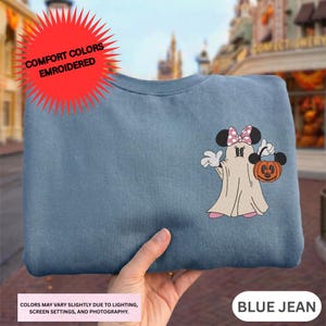 A hand holding a blue t-shirt with a Minnie Mouse and a ghost design, with a red starburst logo above the shirt.