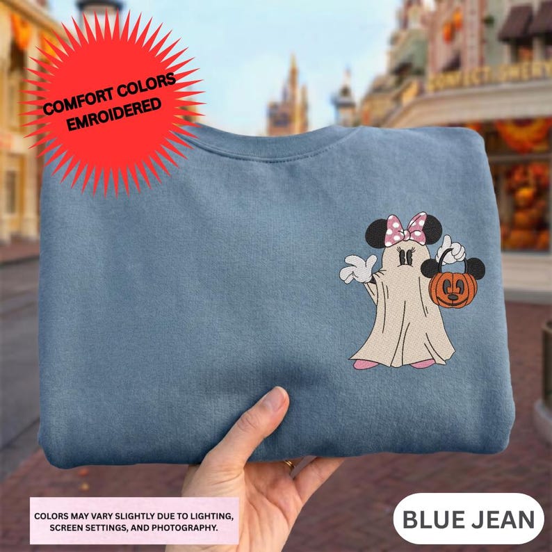 A hand holding a blue t-shirt with a Minnie Mouse and a ghost design, with a red starburst logo above the shirt.
