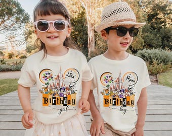 May include: Two cream-colored short-sleeve t-shirts featuring a graphic of a castle, Disney characters, and balloons. One shirt has the text "Birthday girl" with the name "Lisa," and the other has "Birthday boy" with the name "Ryan" printed in black.