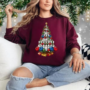 A woman wearing a burgundy sweater with a Christmas tree design on it, sitting on a couch with Christmas decorations in the background.