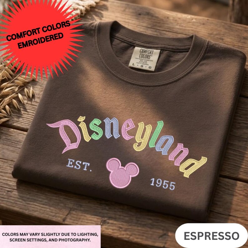 A brown t-shirt with the text "Disneyland" and a Mickey Mouse logo, along with the text "EST. 1955" and "COLORS MAY VARY SLEEPYLY DUE TO LIGHTING, SCREEN SETTINGS AND PHOTOGRAPHY."