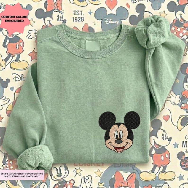 A green long-sleeved t-shirt with a Mickey Mouse embroidered on the front, placed on a background with Disney-themed patterns.