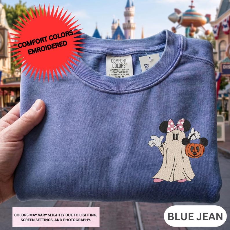 A hand holding a blue t-shirt with a Minnie Mouse and a ghost printed on it, with a castle visible in the background.
