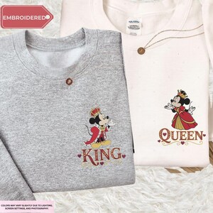 two sweatshirts with a Disney character design, specifically Mickey Mouse. The sweatshirt on the left has the character King and the word "Queen" written on it, while the sweatshirt on the right has the character Queen and the word "King" written on it.