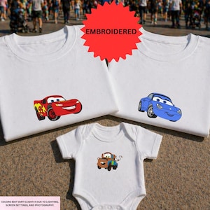 a collection of white t-shirts with colorful cartoon car designs, including a red car, a blue car, and a brown car. The t-shirts are displayed on a carpeted surface, and there is a red starburst logo in the top right corner.