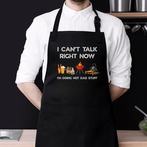 May include: Black apron with white text that reads "I CAN'T TALK RIGHT NOW, I'M DOING HOT DAD STUFF." The apron features illustrations of beer, a truck, a grill, and a lawnmower. The apron is worn by a person wearing a white shirt.