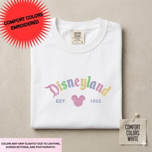 a white t-shirt with a Disneyland logo and text, along with a tag and a label indicating the color options available.
