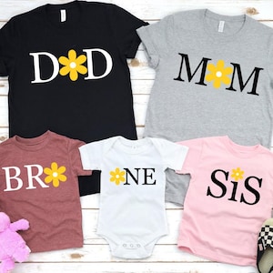 May include: A collection of family matching t-shirts and a onesie. The shirts are black, gray, burgundy, and pink. Each shirt has a word or letters with a yellow flower graphic. The black shirt says "DAD", the gray shirt says "MOM".