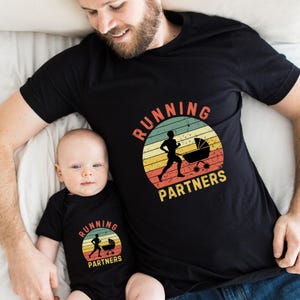 May include: Black t-shirt and onesie set with a retro design. The design features a running figure pushing a stroller, set against a striped sunset background. The text "RUNNING PARTNERS" is arched above and below the image.