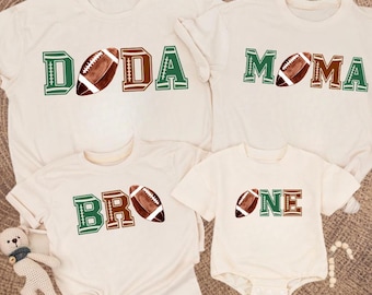 May include: Four cream-colored t-shirts and a onesie with football-themed designs. The shirts read "DODA", "MOMA", and "BRO", each with a football graphic. The onesie says "ONE" with a football. The text is in green and brown.