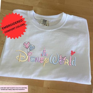 A white t-shirt with the text "DisneyWorld" embroidered on it, along with various Disney-themed elements such as Mickey Mouse, Minnie Mouse, and a castle.