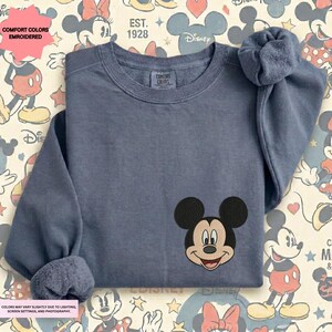 A gray long-sleeved t-shirt with a Mickey Mouse embroidered on the front, placed on a background with Disney-themed graphics.