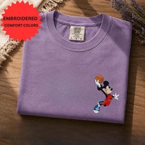 a purple t-shirt with a cartoon character, specifically Mickey Mouse, depicted in a basketball pose. The t-shirt is placed on a wooden surface, and there is a red starburst sign in the top left corner.
