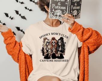 May include: A cream-colored t-shirt with the text "Spooky Mom's Club" and the phrase "Caffeine Required." The shirt features a graphic of three skeleton women holding coffee cups. The person is wearing an orange sweater and holding a book that says "Spells & Potions."