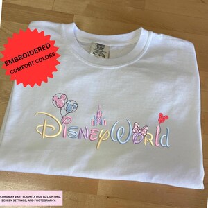 A white t-shirt with the text "DisneyWorld" embroidered on it, along with various Disney-themed elements such as Mickey Mouse, Minnie Mouse, and a castle.
