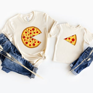 May include: Two cream-colored t-shirts with pizza slice graphics. One shirt features a large pizza with missing slices, and the other has a single slice. Blue jeans are next to the shirts. The pizza design includes red pepperoni and brown mushrooms.