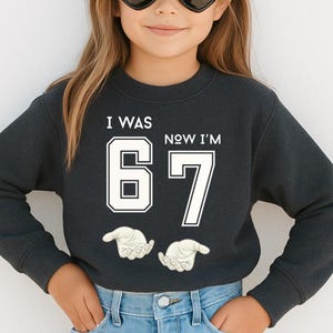A young girl wearing a black sweatshirt with the text "I WAS 67 NOW I'M" printed on it.