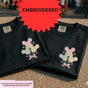 two black t-shirts with a Mickey Mouse design, including a balloon and stars, placed on a surface.